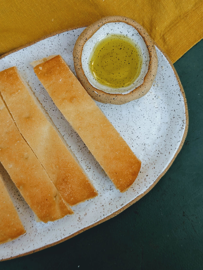 Speckled plate with four breadstick slices and a small bowl of olive oil on a green table and yellow napkin