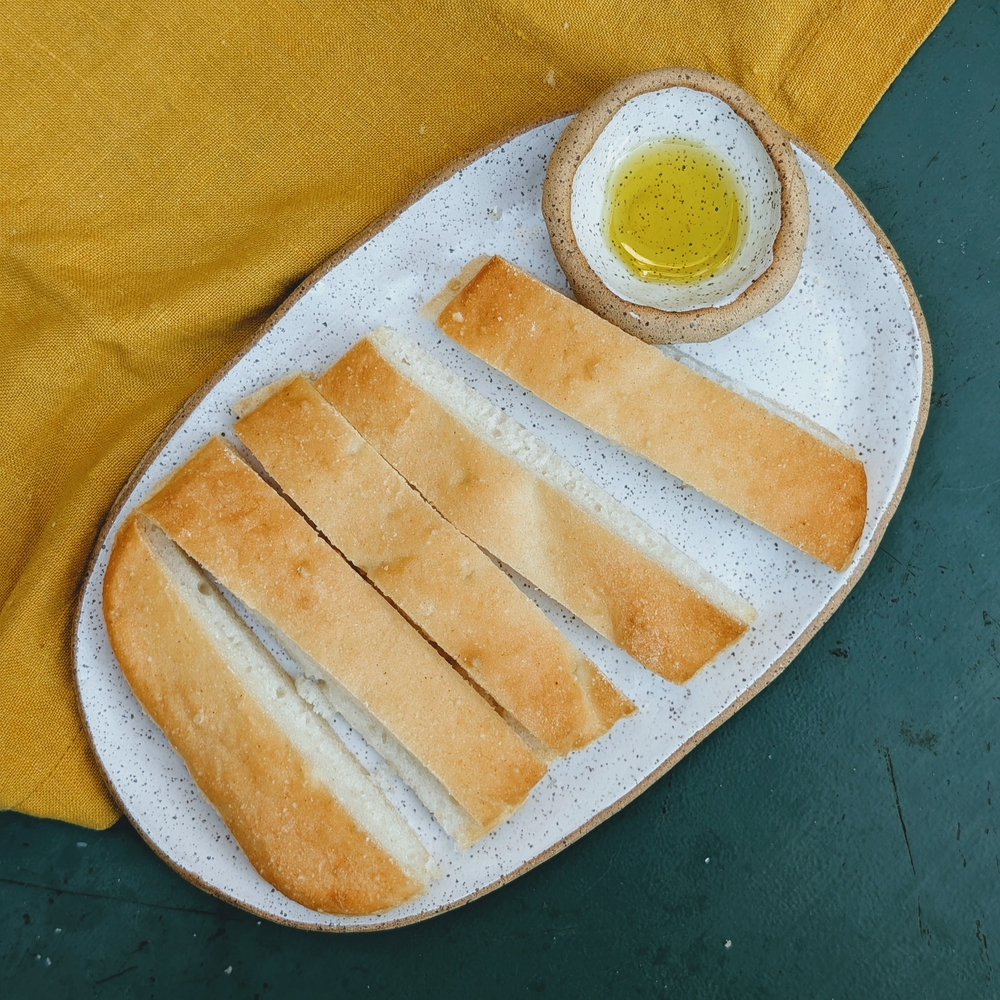 Sliced baguette arranged on a speckled oval plate with a small bowl of olive oil, mustard-yellow napkin at top-left.