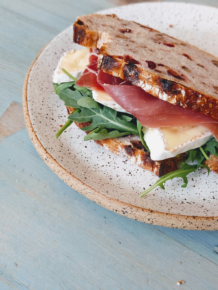 Sandwich with arugula, prosciutto, and cheese on a speckled plate with a light blue background