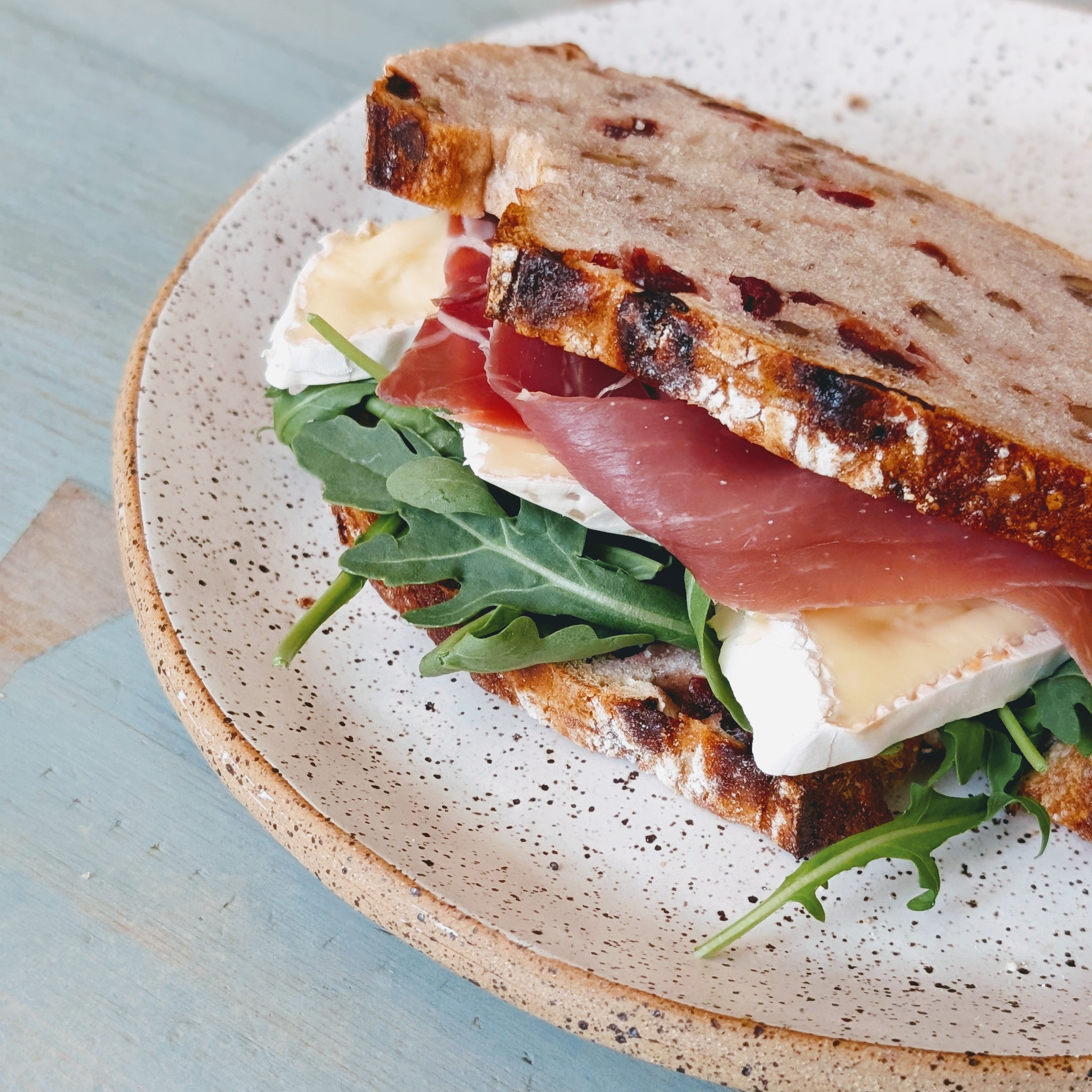 Toasted cranberry-walnut bread sandwich with prosciutto, brie cheese, and arugula on a speckled ceramic plate.