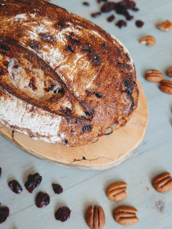 Crusty artisan cranberry-pecan loaf on a wooden board, with scattered pecans and dried cranberries.