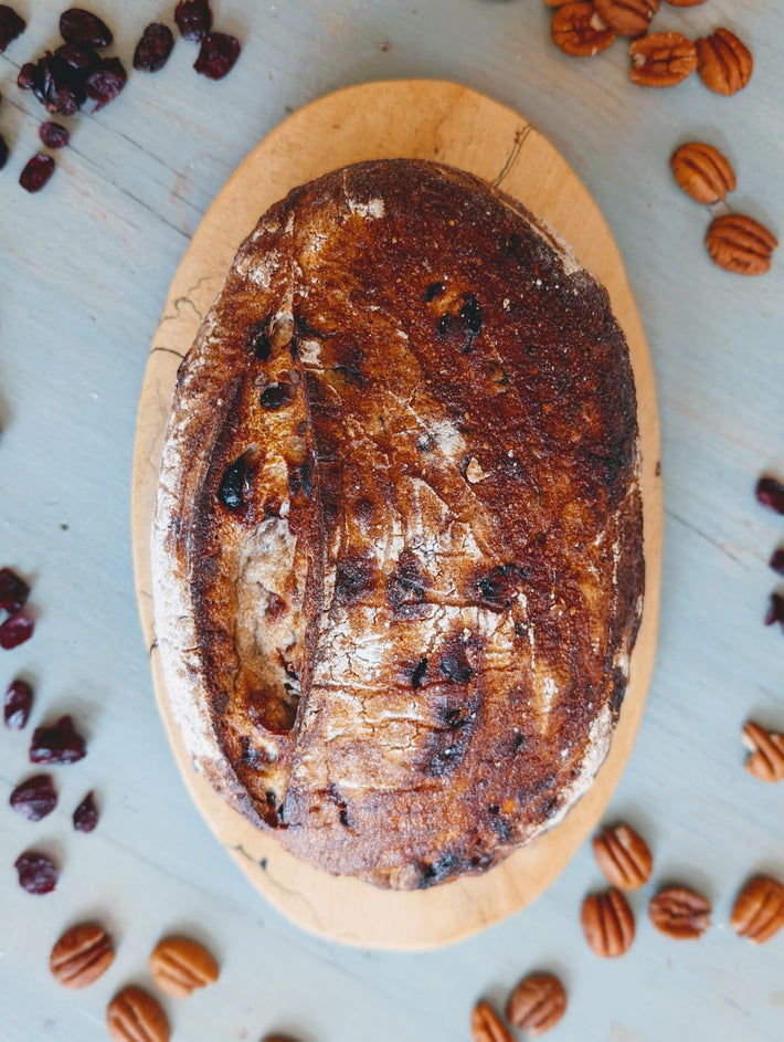 Rustic cranberry-pecan sourdough loaf on an oval wooden board, dried cranberries and pecans scattered on a pale blue tabletop
