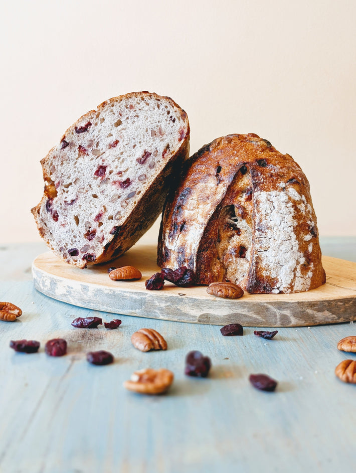 Cranberry-pecan sourdough, one half sliced to show crumb, on a wooden board with scattered pecans and dried cranberries.