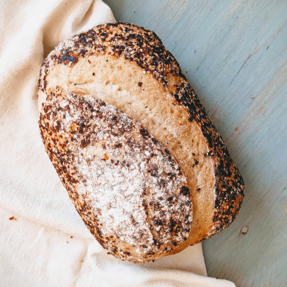 Oval seeded artisanal loaf with flour-dusted top resting on a white cloth on a pale blue wooden surface.