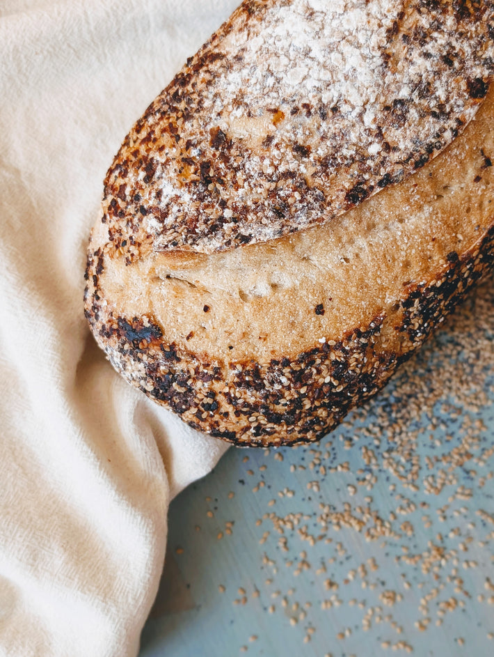 Crusty seeded sourdough loaf on a white cloth with sesame seeds scattered across a pale blue table.