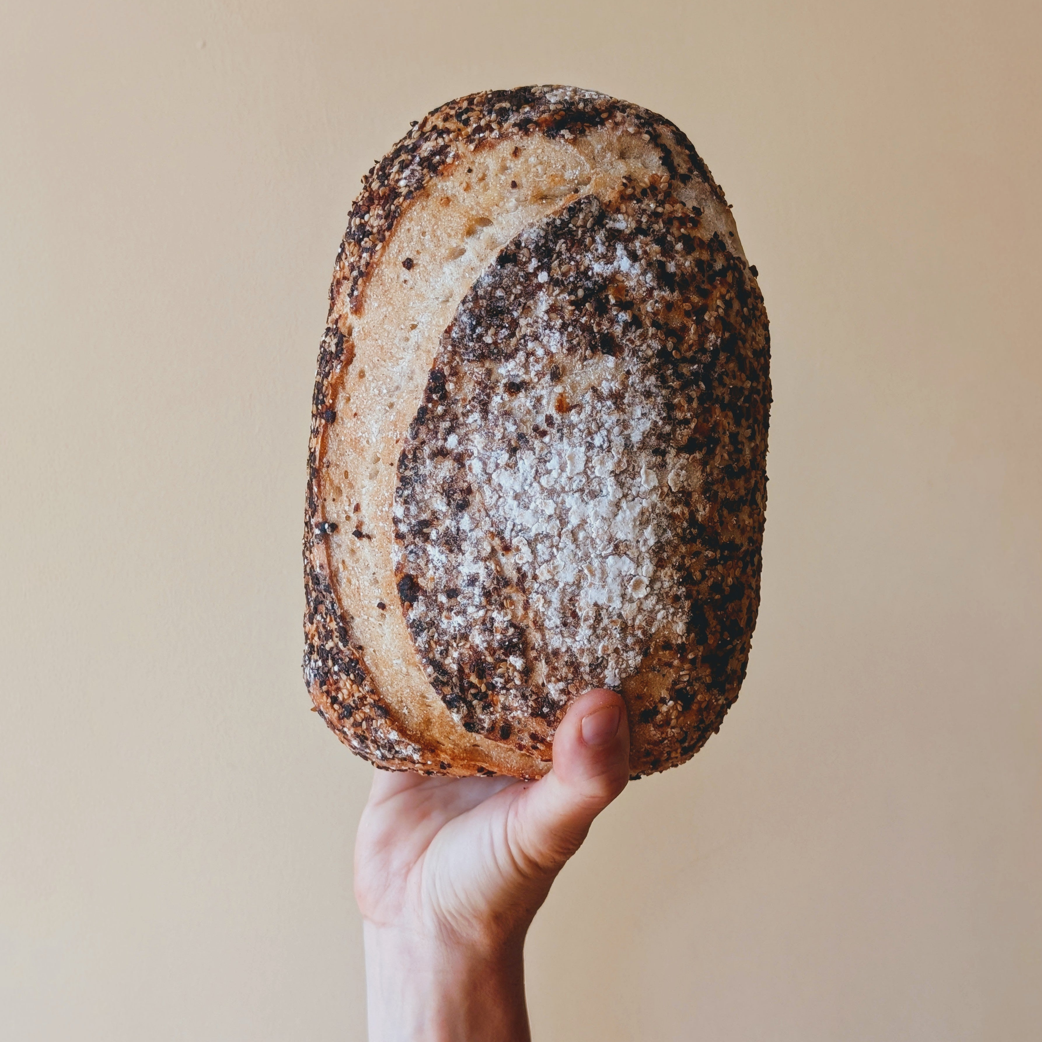 Hand holding a seeded artisan loaf dusted with flour against a beige background.