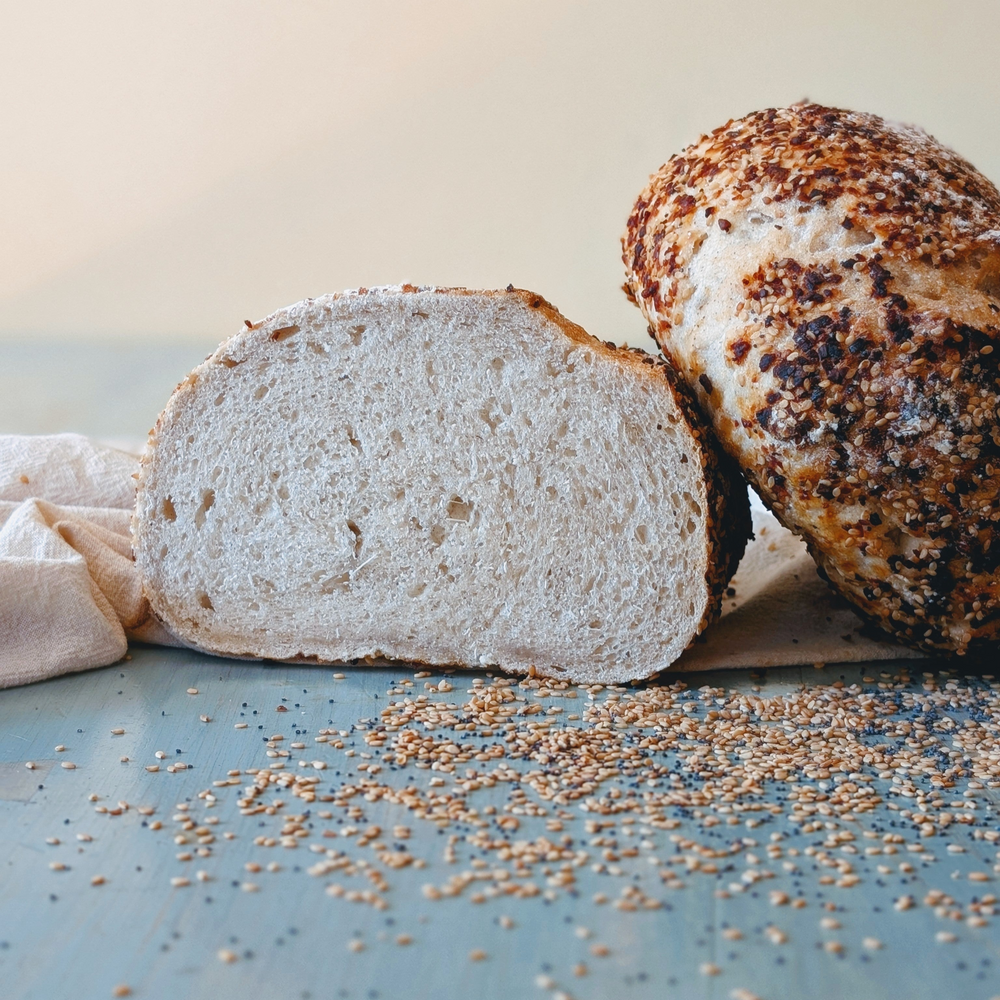 Two loaves of sesame-crusted artisan bread; one sliced to show the crumb, on a blue tabletop with scattered sesame seeds.
