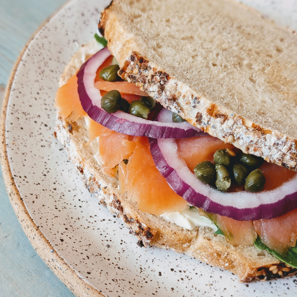 Smoked salmon sandwich with red onion rings, capers and cream cheese on seeded bread served on a speckled plate.
