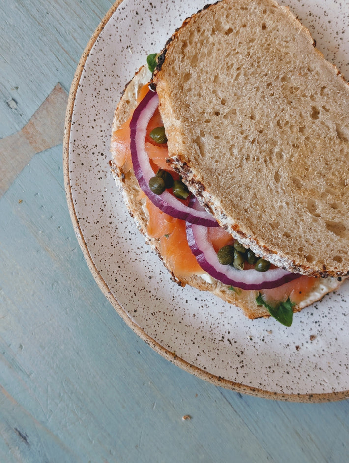 Smoked salmon sandwich with red onion rings and capers on seeded bread, served on a speckled ceramic plate over a blue table.