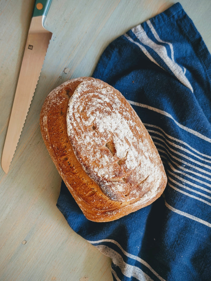 Loaf of crusty artisan bread dusted with flour on a blue striped kitchen towel beside a serrated bread knife.