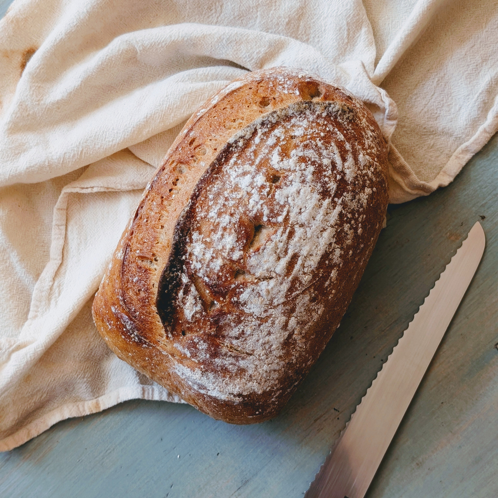 Crusty oval artisan loaf dusted with flour on a cloth, beside a serrated bread knife on a wooden surface