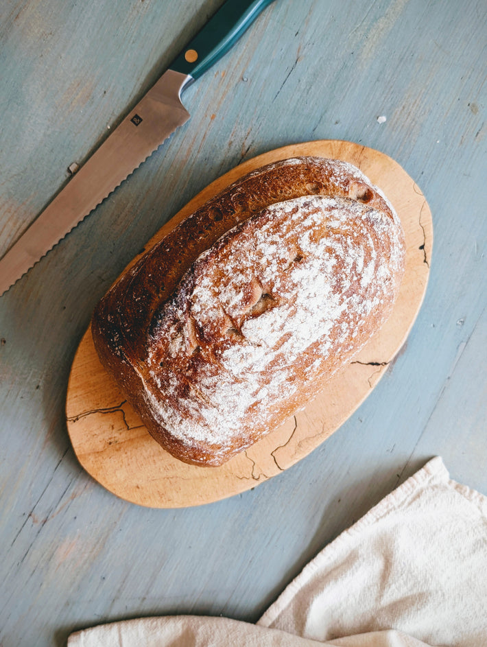 Rustic loaf of crusty bread dusted with flour on a wooden cutting board beside a serrated knife and kitchen towel.