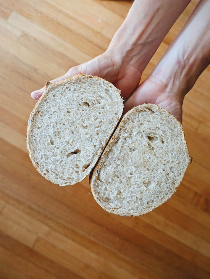 Two halves of a sliced round loaf held palm-up over a wooden floor, showing crumb texture and crust.