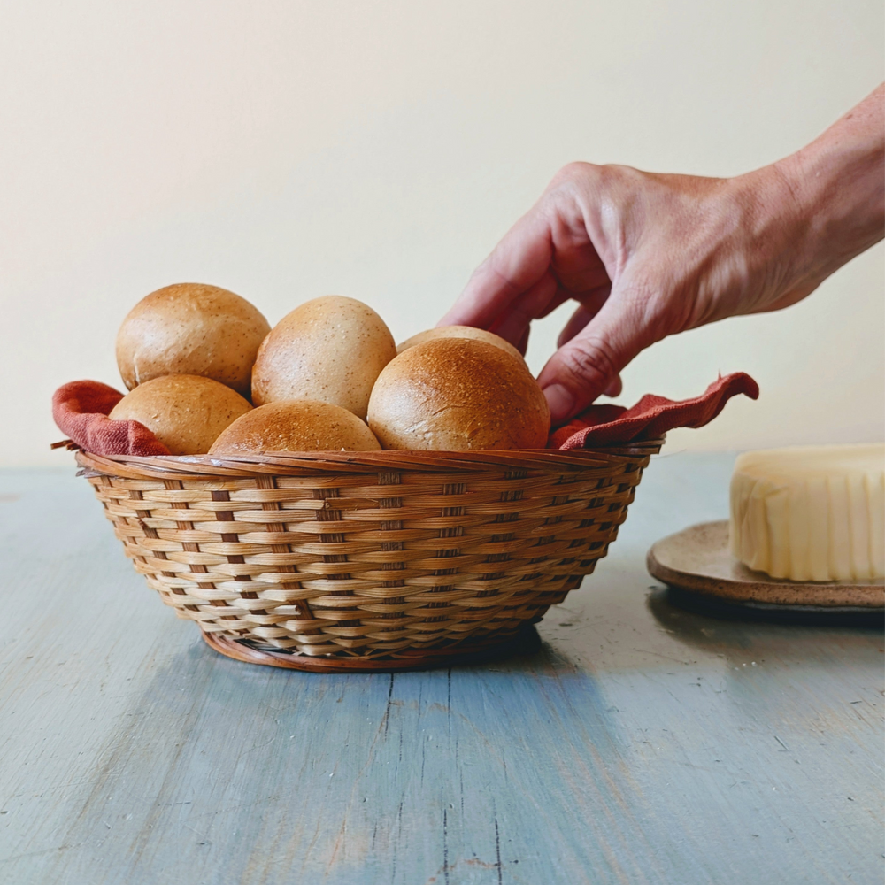 Wicker basket of round bread rolls on a blue wooden table, hand reaching for a roll, butter on a plate at right.
