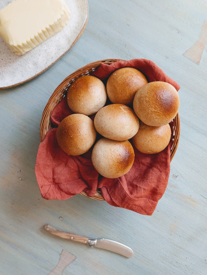 Wicker basket with bread rolls on a red cloth, next to a block of butter and knife on a light blue surface.