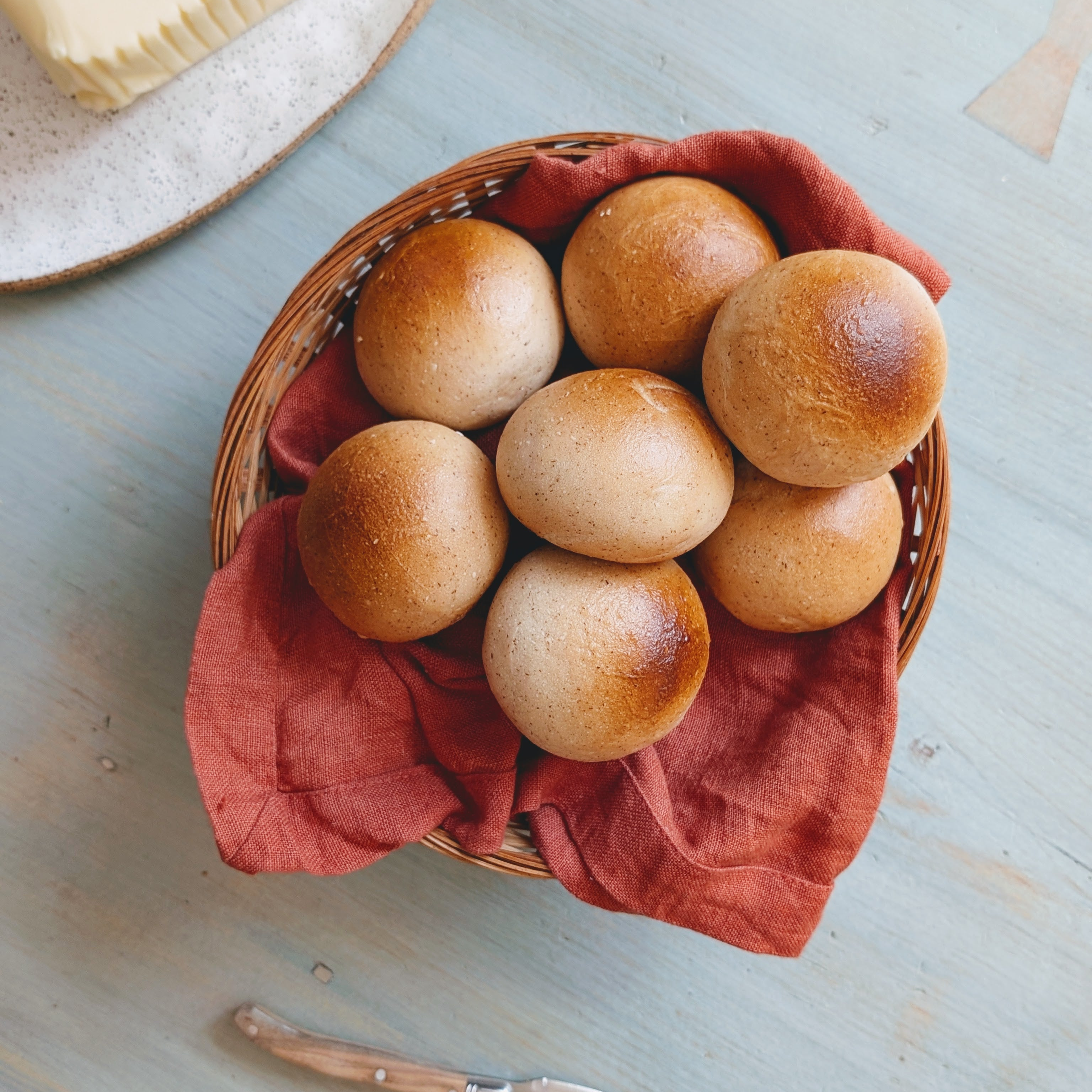 Wicker basket lined with a red cloth holding seven round golden-brown bread rolls on a pale blue table