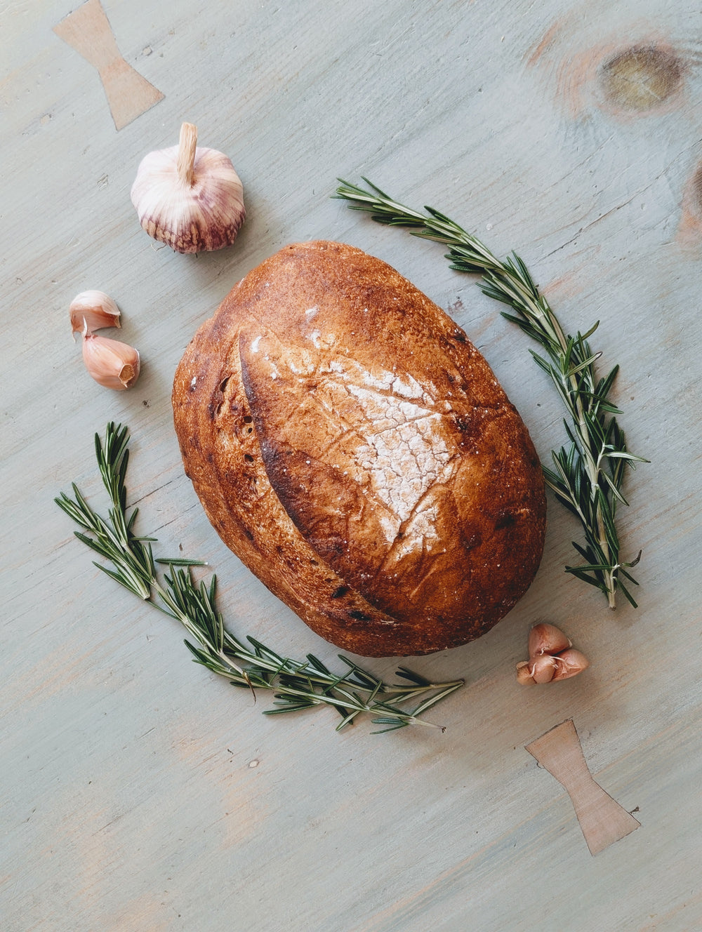 Round rustic sourdough loaf with garlic cloves and rosemary sprigs on painted wooden table
