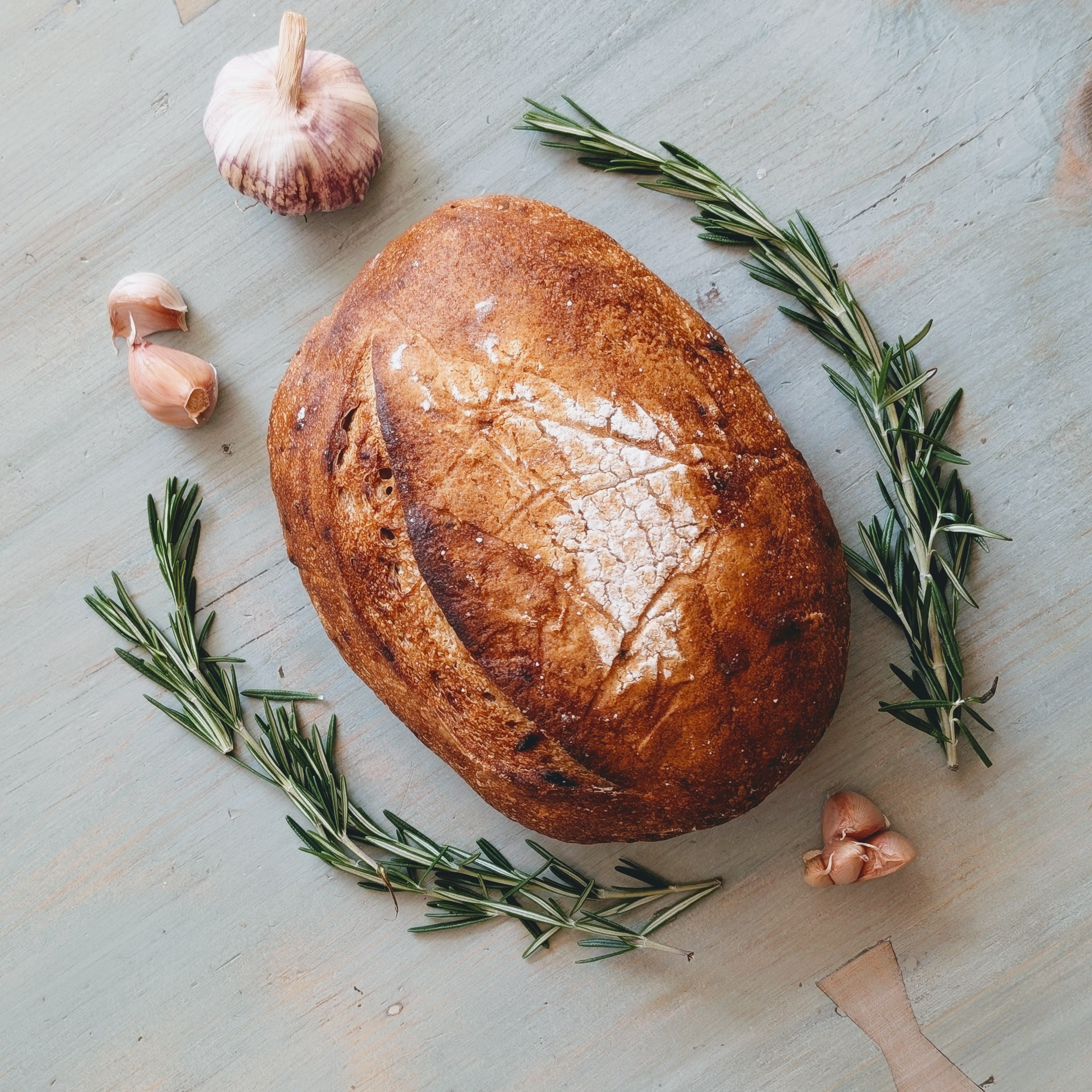 Rustic sourdough loaf with rosemary sprigs and garlic cloves arranged on a pale wooden surface