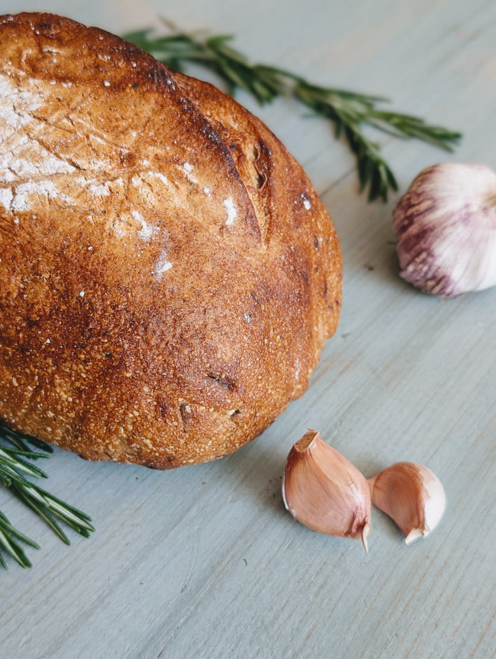 Sourdough loaf with golden crust, two garlic cloves and rosemary sprigs on a pale wooden surface.