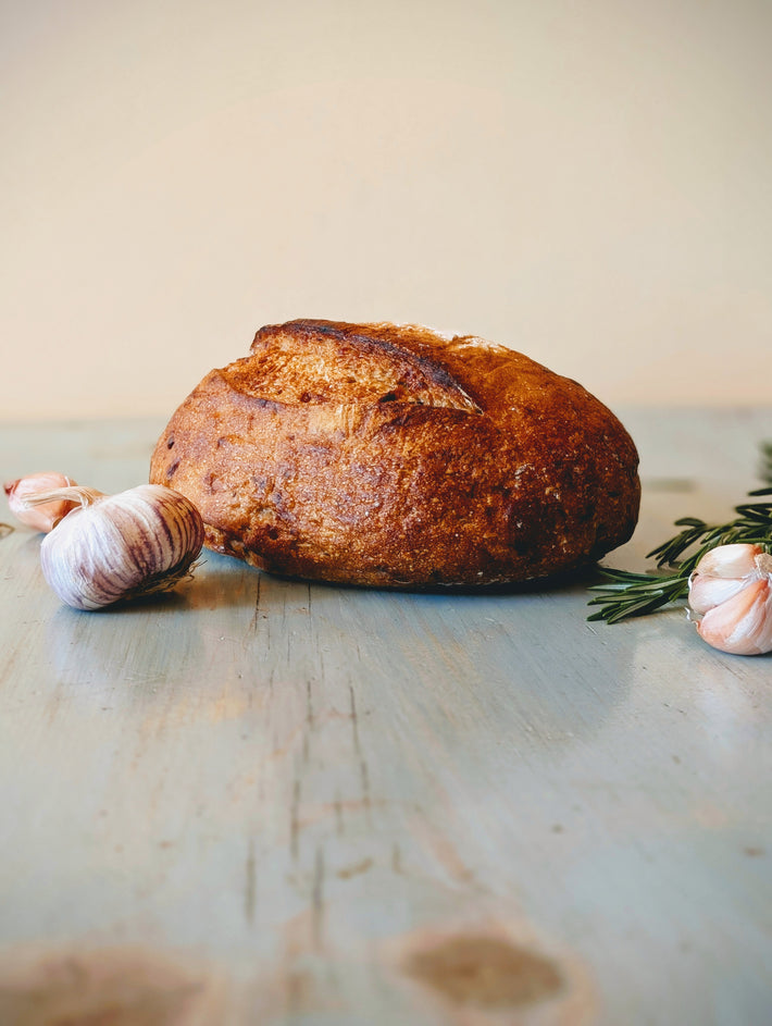 Round crusty sourdough loaf on a painted wooden table with garlic bulbs and rosemary sprigs.