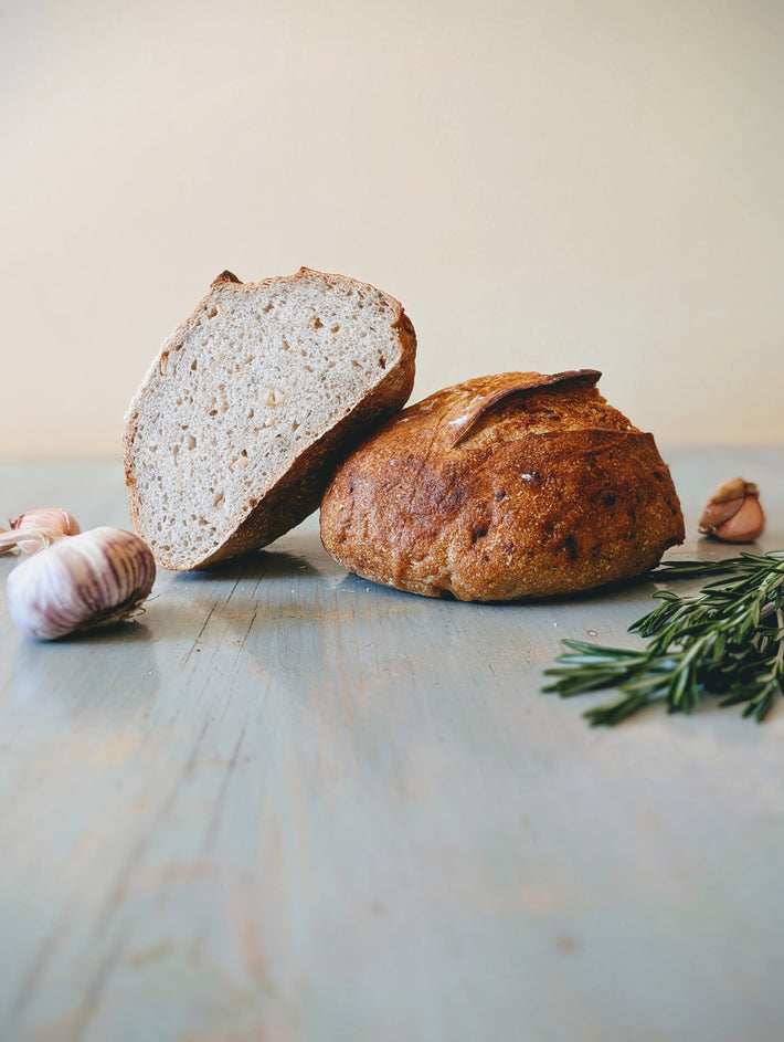 Two rustic sourdough loaves on a blue wooden table, one sliced to show crumb, garlic bulbs and rosemary sprigs nearby.