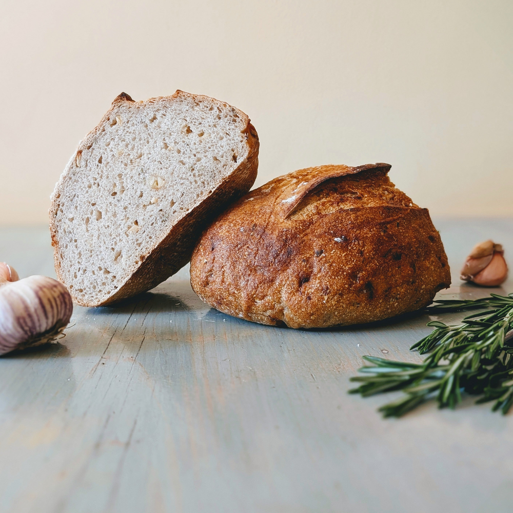Rustic round loaf, one half cut to reveal the crumb, on a blue wooden table with garlic cloves and a rosemary sprig.