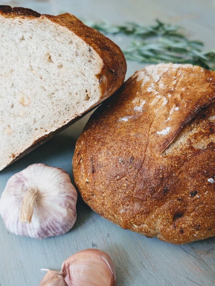 Two crusty loaves (one halved) with a garlic bulb and a garlic clove on a light blue surface.