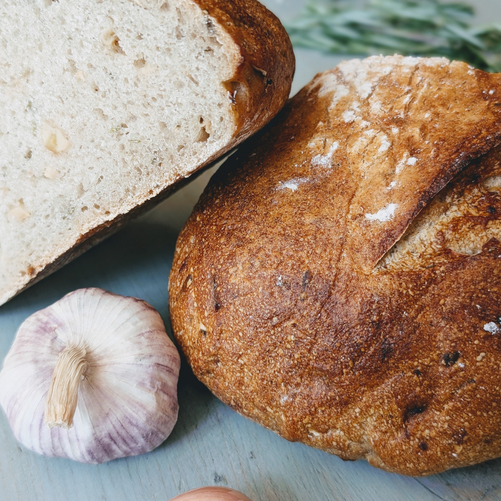 Crusty round artisanal loaf with a sliced half, garlic cloves nearby on a pale blue surface.