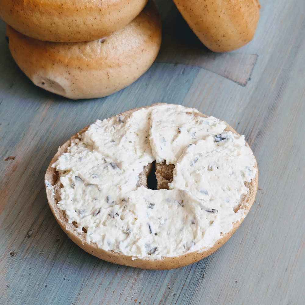 Halved toasted bagel spread with herb cream cheese on a blue wooden table, with stacked bagels in the background.