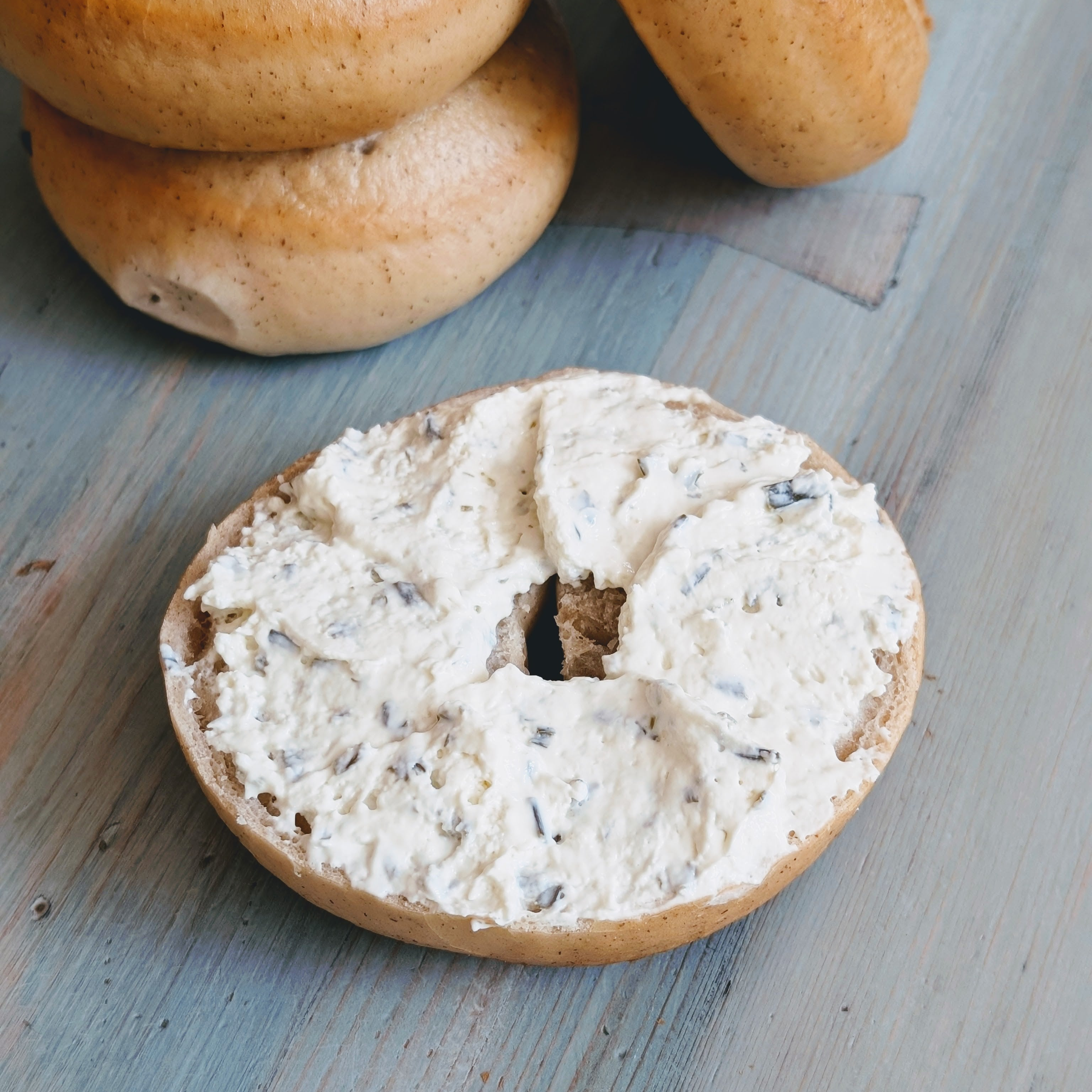 Halved toasted bagel spread with herb cream cheese on a blue wooden table, with stacked bagels in the background.
