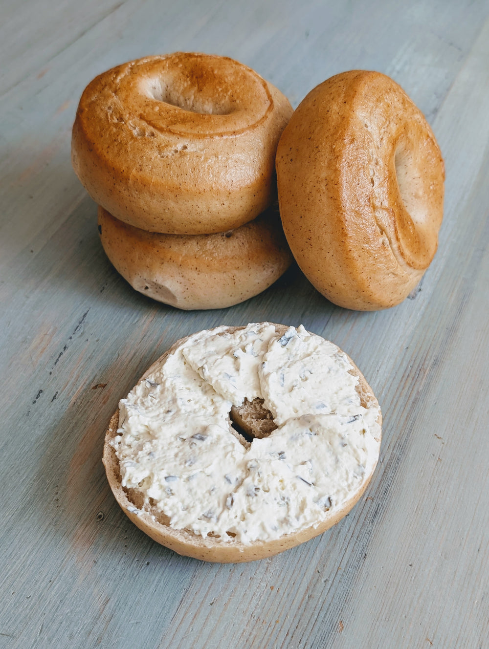 Front view of gluten-free plain bagels showcasing one sliced bagel spread with cream cheese on a light wood surface