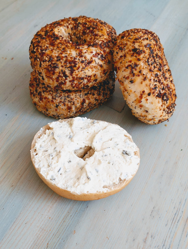 Three everything bagels, one sliced and spread with herbed cream cheese, on a light blue wooden surface.
