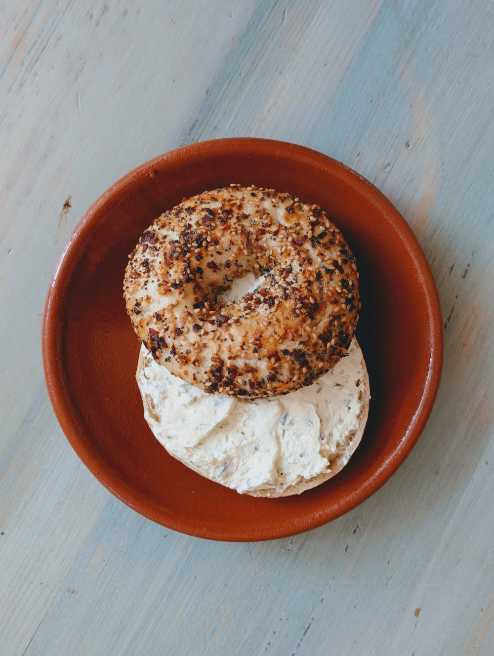 Top view of a gluten-free everything bagel halved and spread with cream cheese on a small terracotta plate on a light blue wooden surface