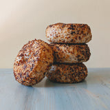 Side view of four gluten-free, slow-fermented everything bagels stacked to show seeded golden crust on a pale blue wooden surface