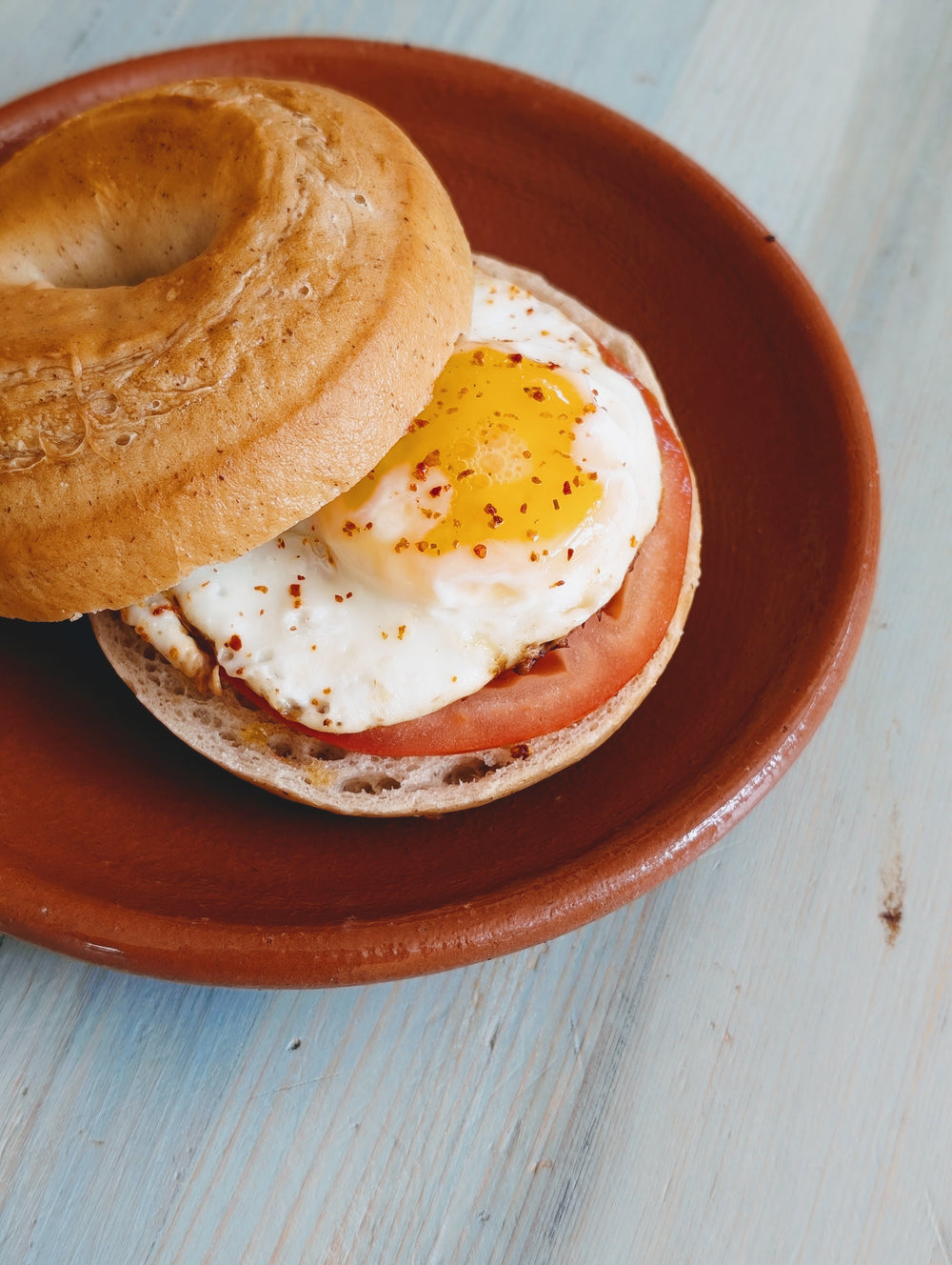 Toasted bagel sandwich with fried egg, tomato slice and red pepper flakes on a brown plate over light blue wood table.