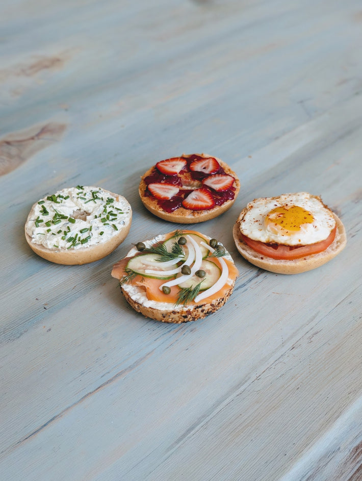 Four bagels on a wooden surface, topped with cream cheese, strawberry jam, smoked salmon, and a fried egg.