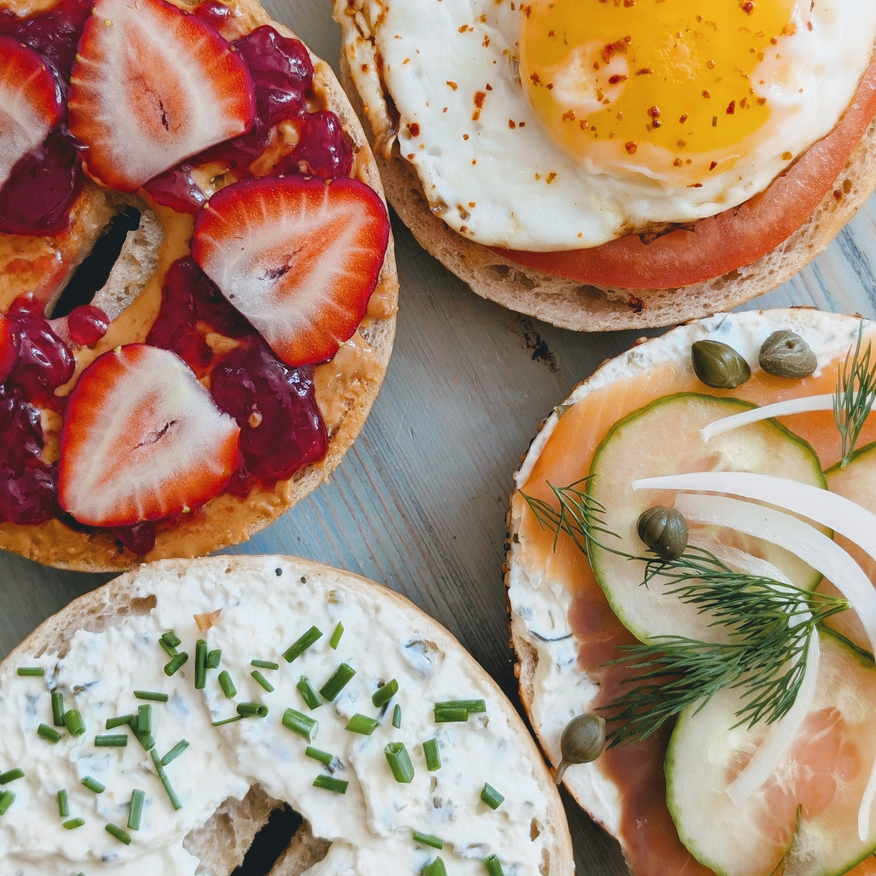Four open bagels: peanut butter, jam and strawberries; fried egg; herbed cream cheese; smoked salmon with cucumber.