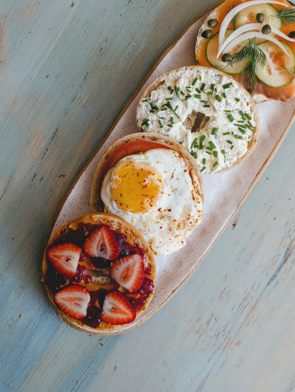 Overhead view of gluten-free bagels topped with strawberries and jam, a fried egg, chive cream cheese, and smoked salmon on a speckled plate