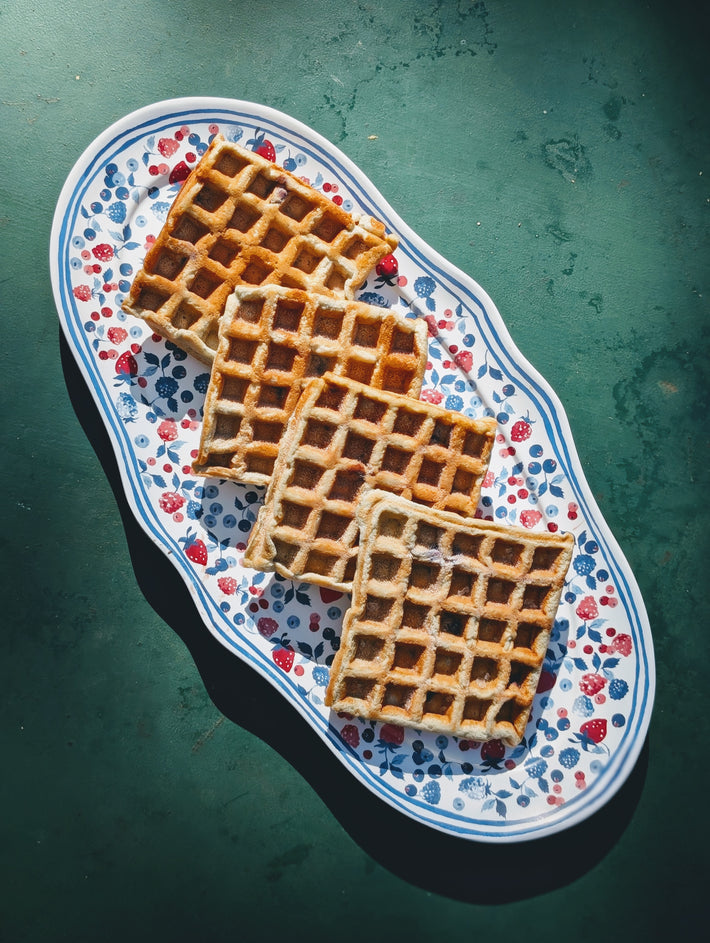 Four golden Belgian-style waffles stacked on a floral oval platter with a blue rim set on a dark green surface