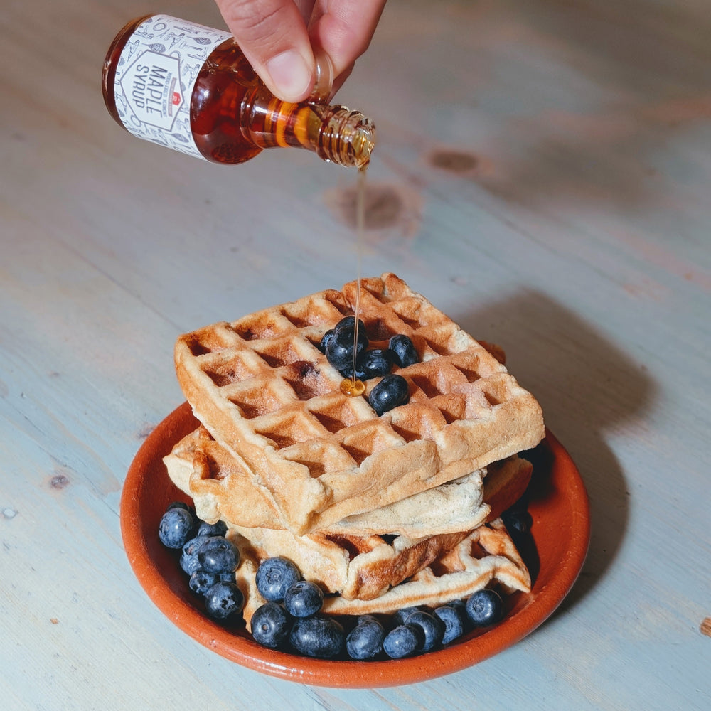 Stack of waffles with blueberries on a small plate while syrup is poured from a glass bottle