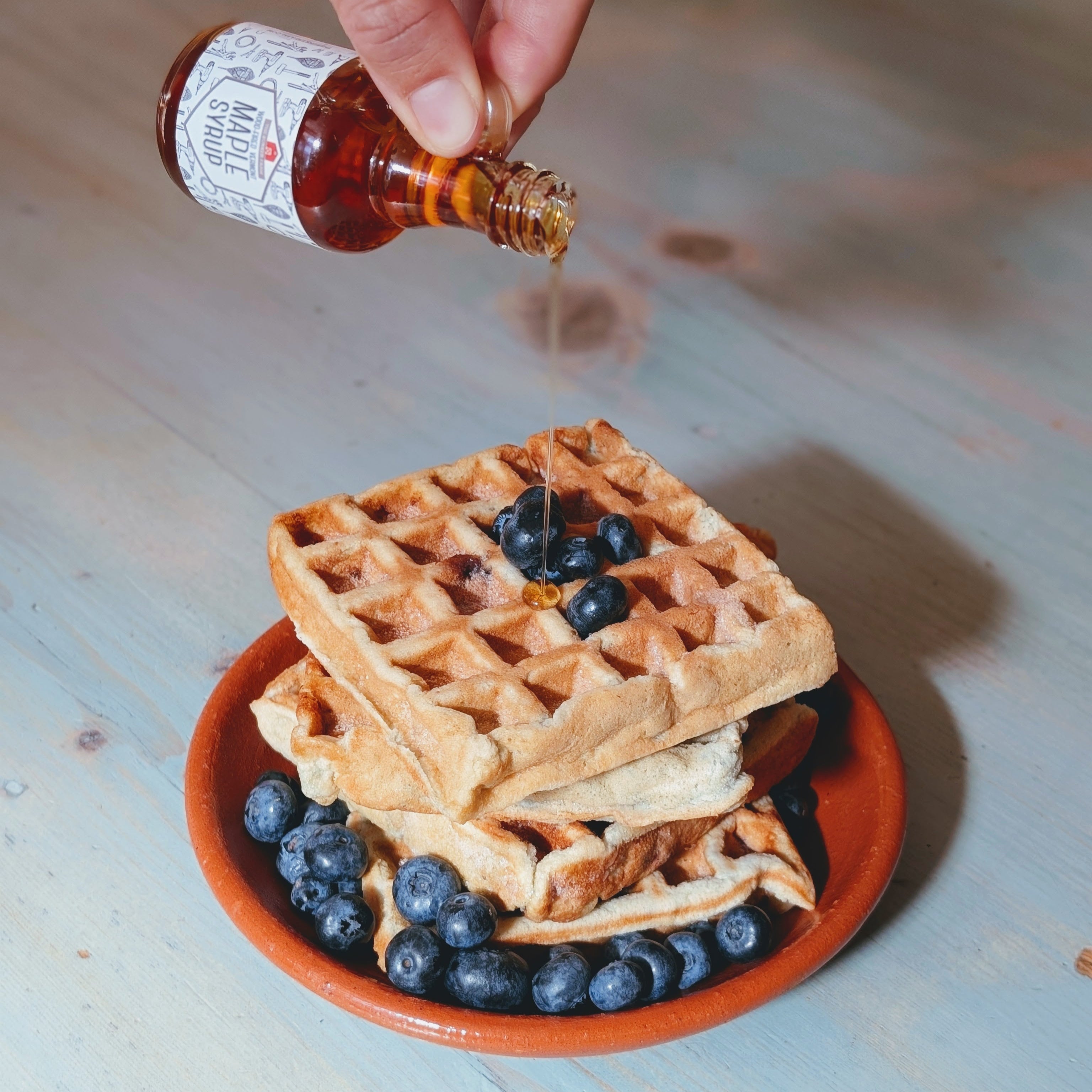Stack of waffles with blueberries on a small plate while syrup is poured from a glass bottle
