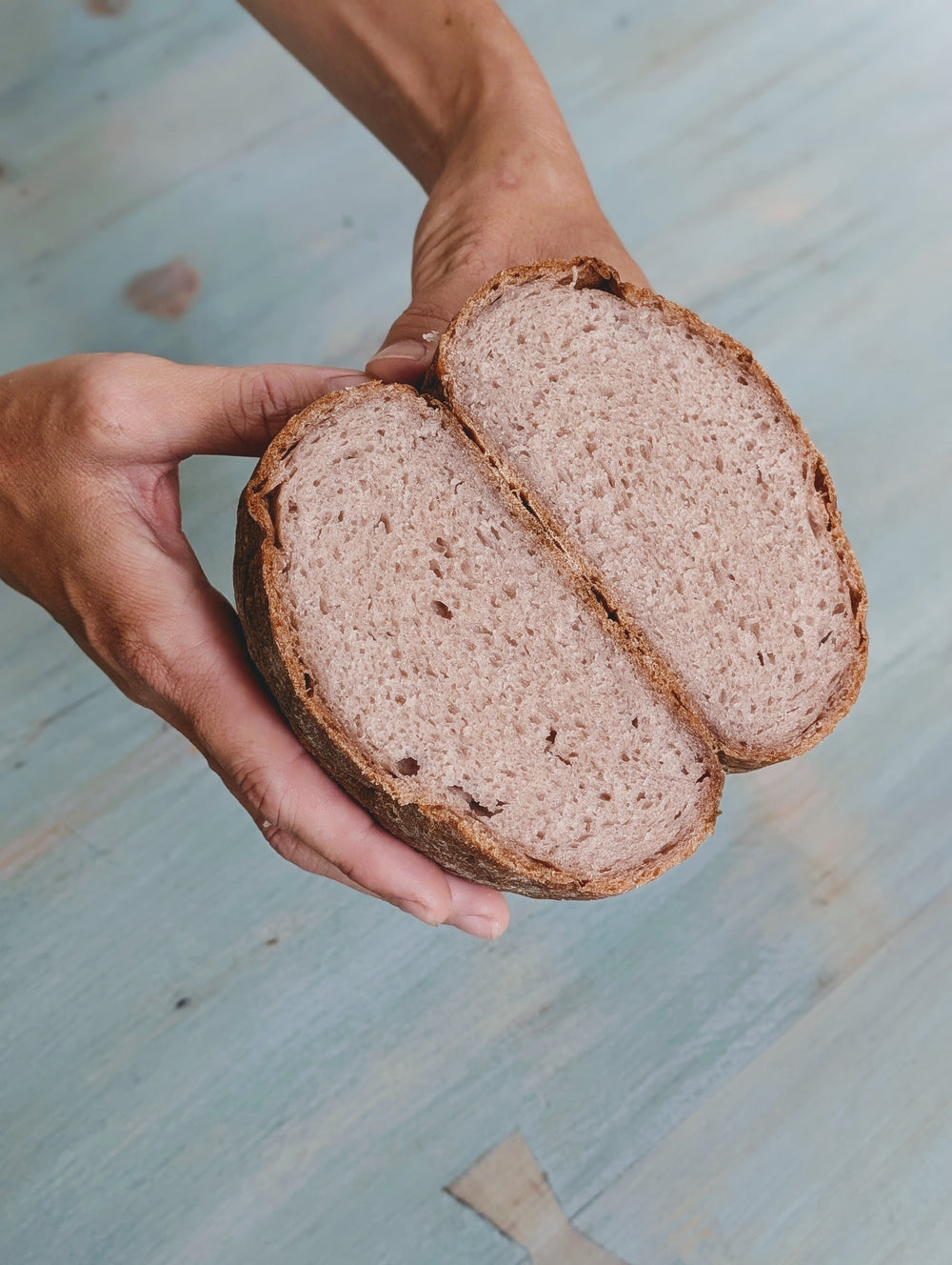 Hands holding a halved round sourdough loaf showing open crumb and crust over a blue wood surface