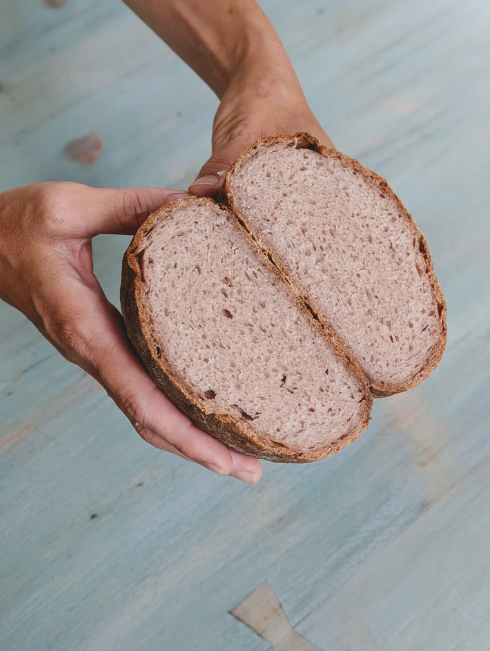 Hands holding a halved round sourdough loaf showing open crumb and crust over a blue wood surface