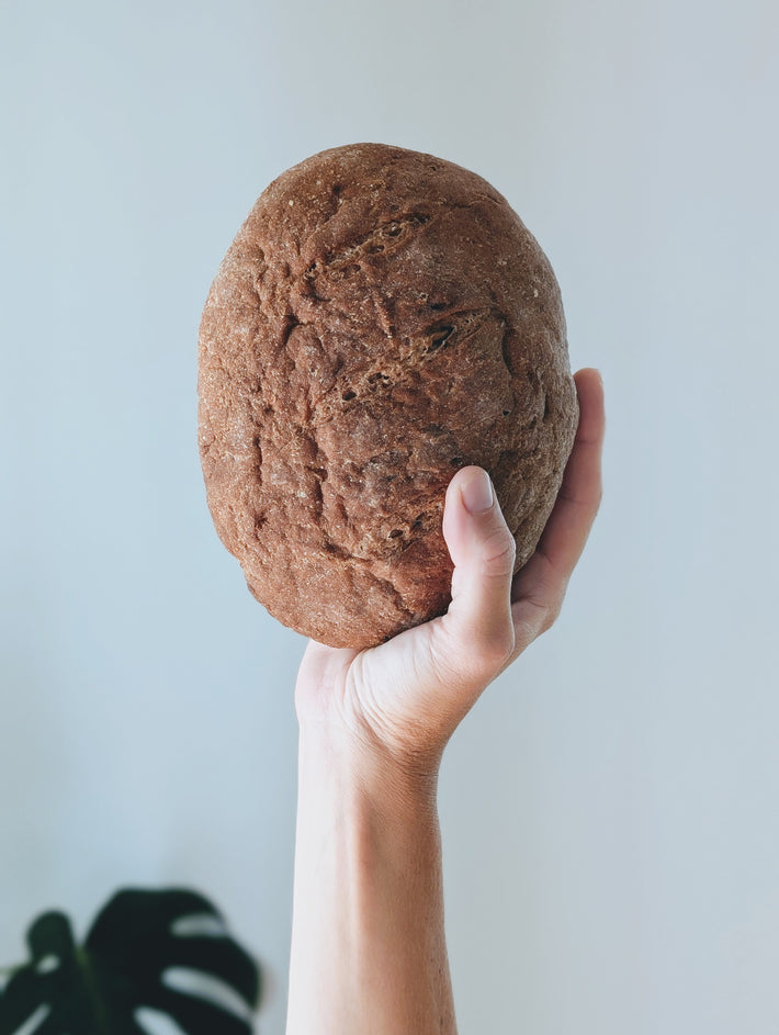 A hand holds a round rustic brown country loaf against a pale background.
