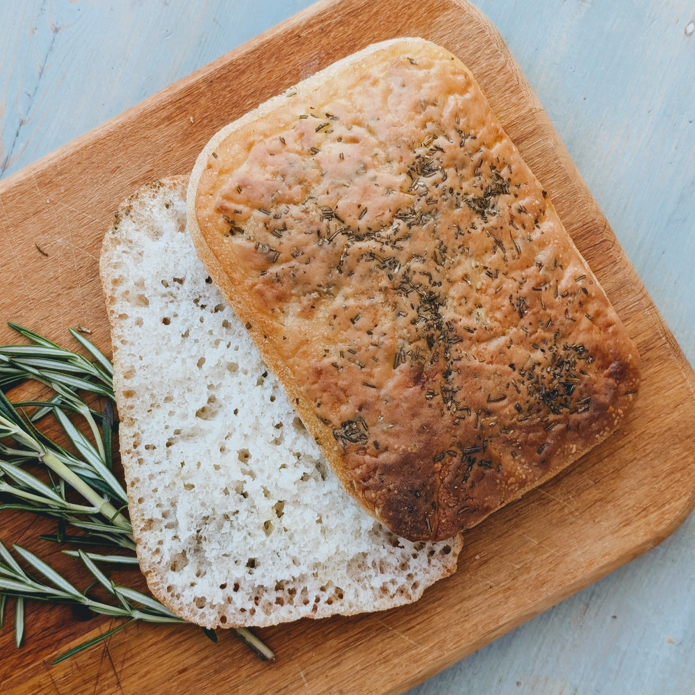 Rectangular rosemary focaccia on a wooden cutting board, one slice flipped to show the airy crumb.
