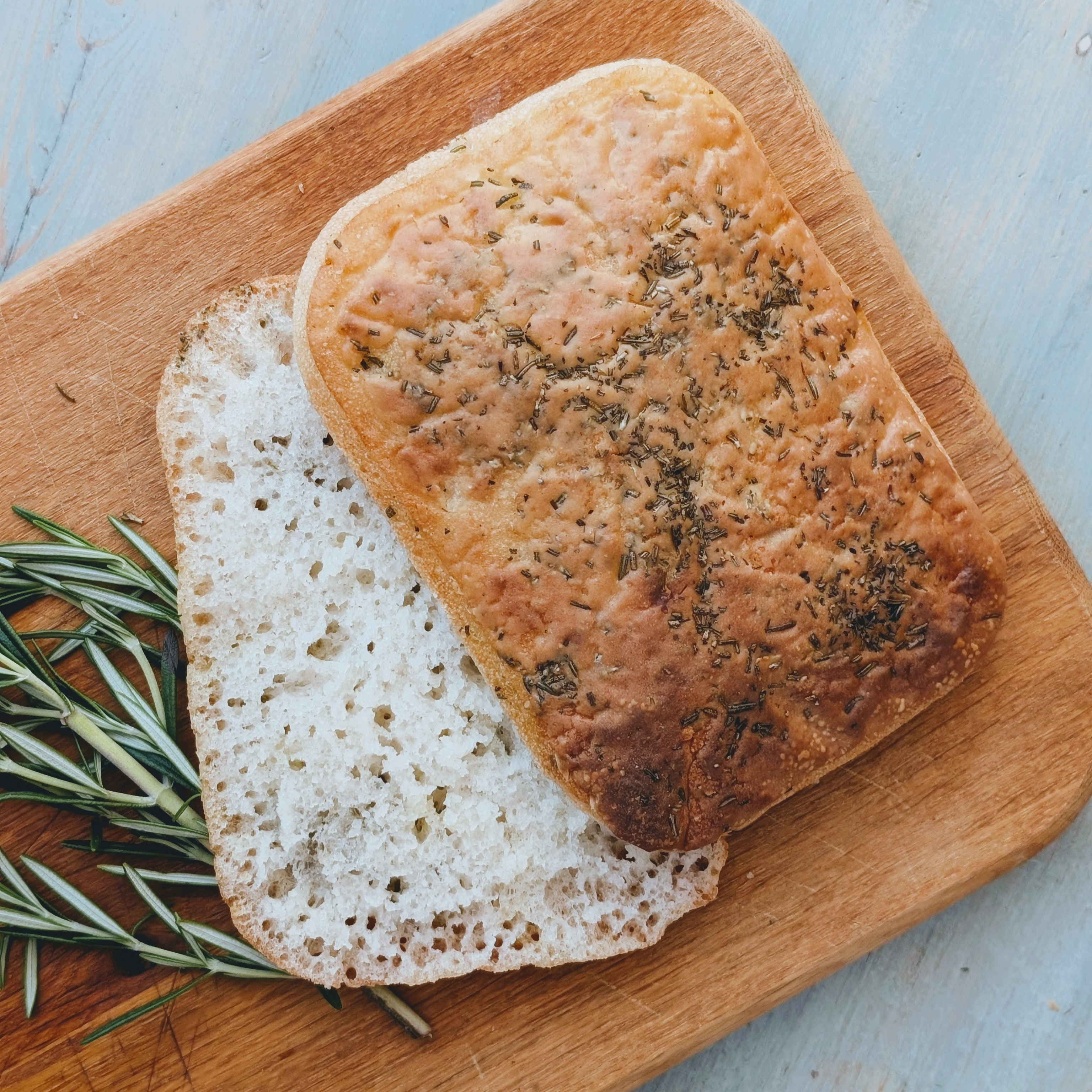 Rectangular rosemary focaccia on a wooden cutting board, one slice flipped to show the airy crumb.