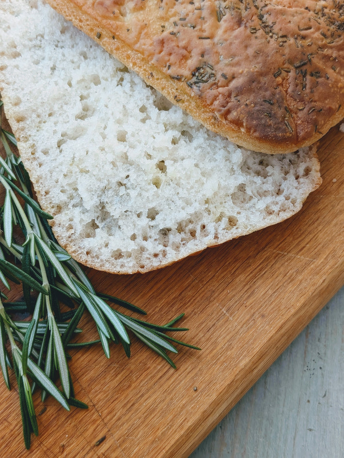 Gluten-free focaccia slice on a wooden board with fresh rosemary sprigs.