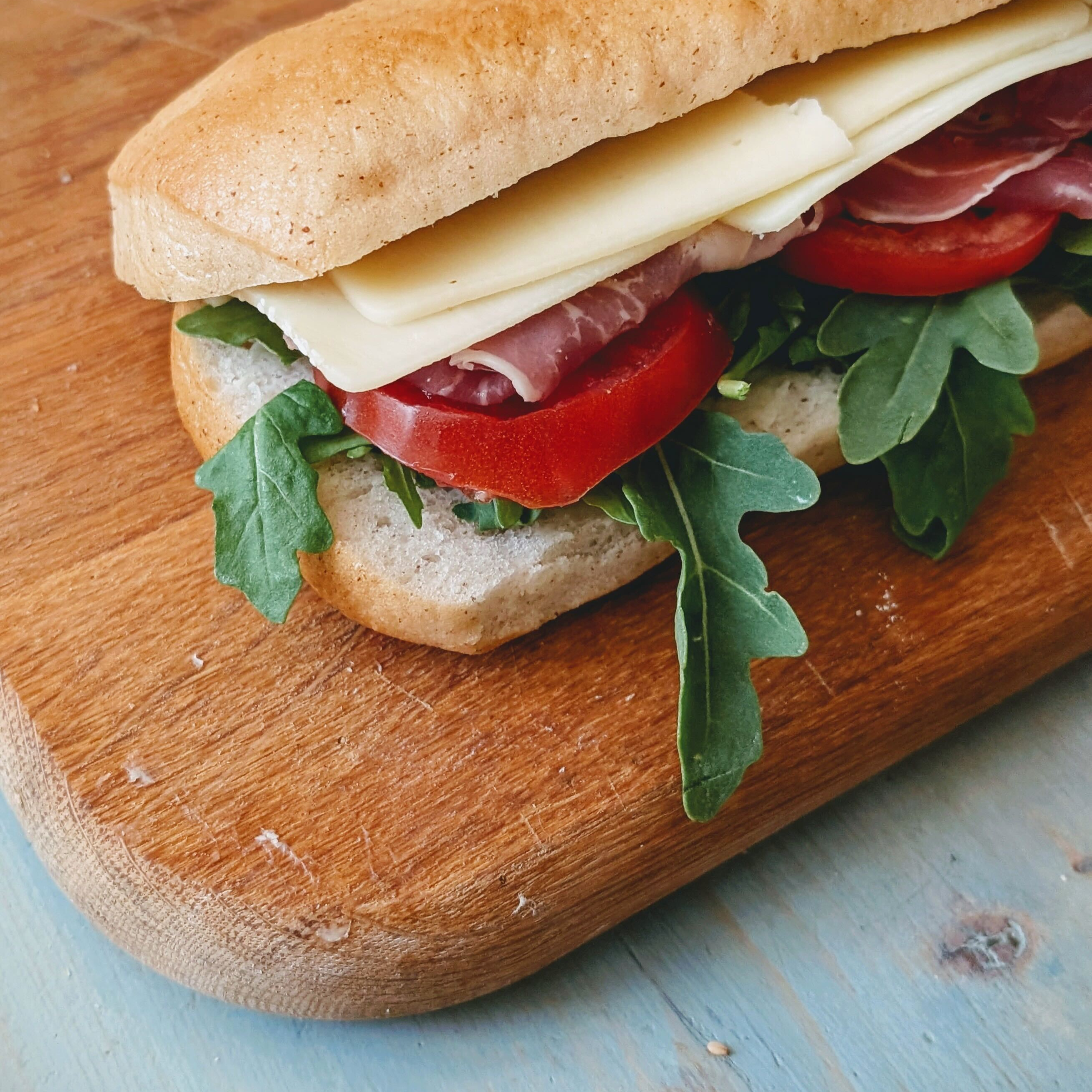 Baguette sandwich with cheese, cured meat, tomato slices and arugula on a wooden cutting board.