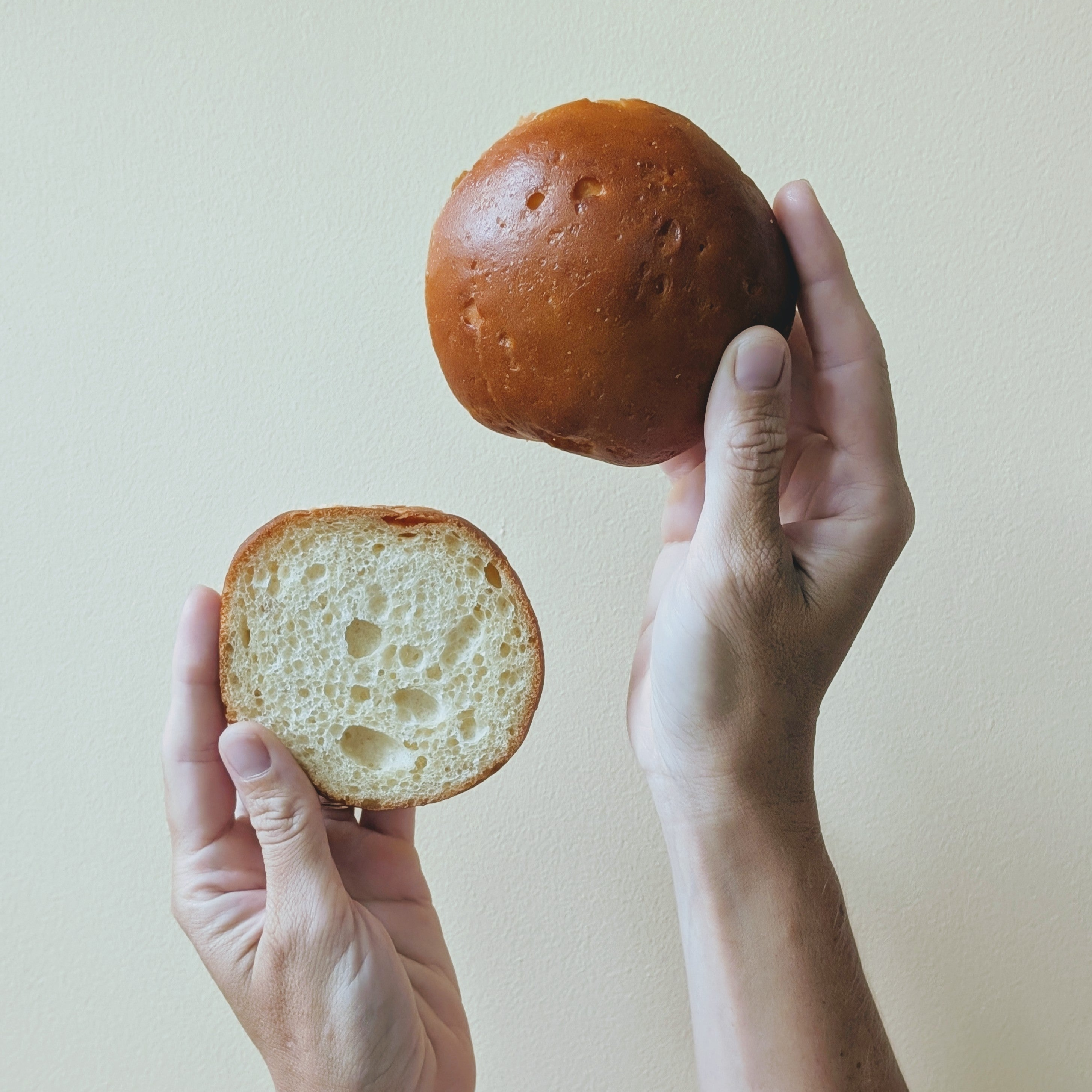 Hands holding a split bun: one hand holds the shiny browned top, the other shows the airy interior crumb.