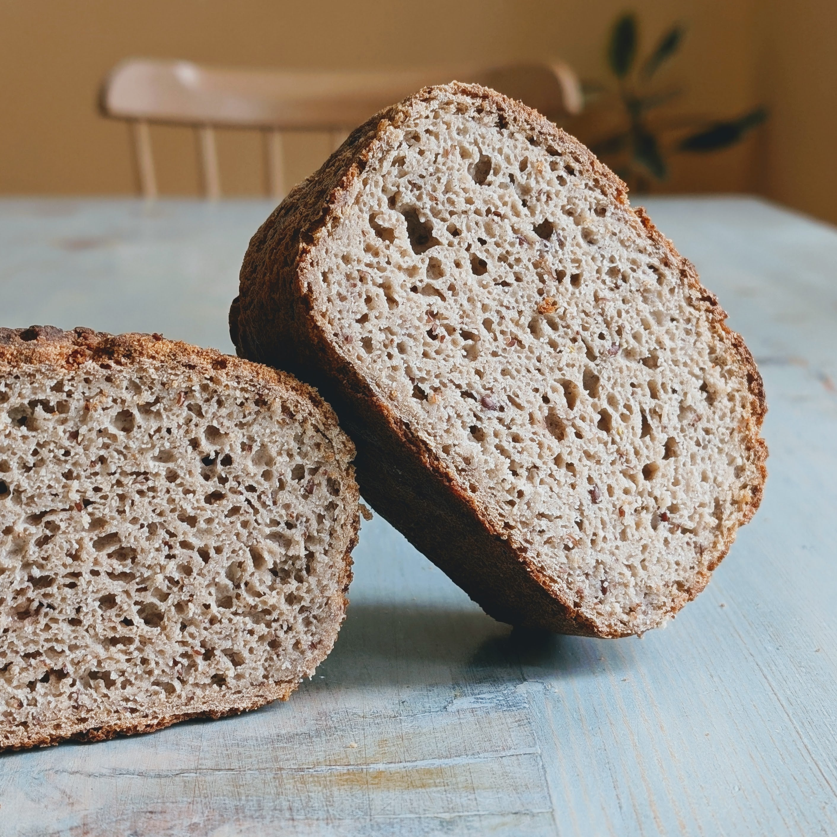 Two halves of rustic whole-grain bread resting on a painted blue wooden table, chair and plant blurred in background.
